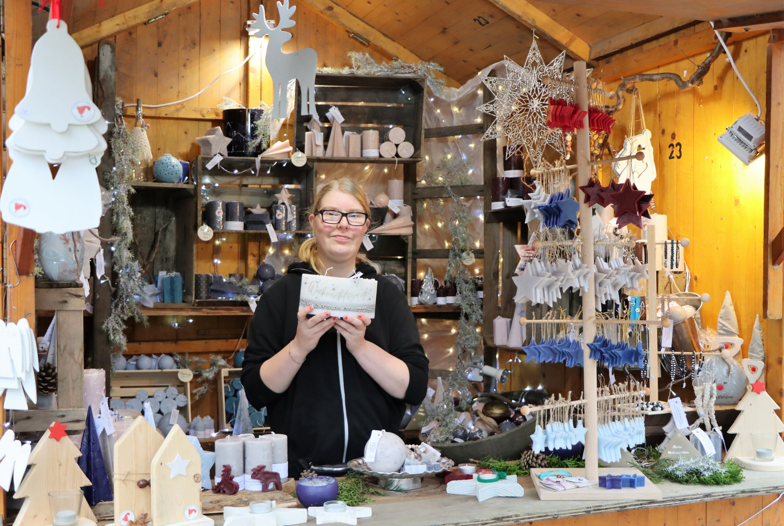 Einen Stand mit einer jungen Frau mit weihnachtlichen gebastelten Geschenken (zum Beispiel Glöckchen mit Bänder, Tannenbaum und Häuser aus Holz, Wachskerzen, usw.) Geschenkartikel von der GPS Werkstatt Jeringhave