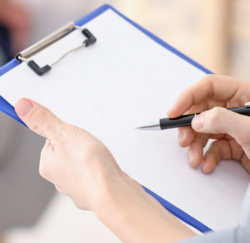 Eine Frau sitzt mit einem Notizbrett mit Blättern und einem Stift. Eine Frau sitzt auch im Hintergrund.