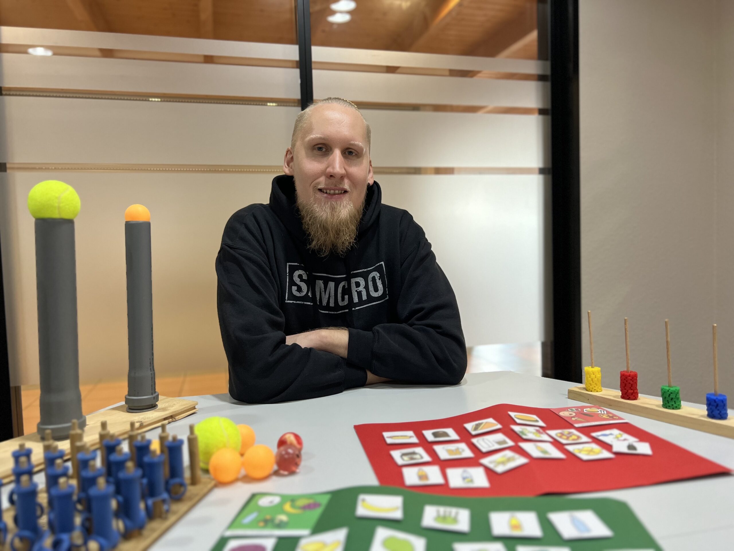 Ein junger Mann sieht an einem Tisch in einem Raum. Er lächelt.