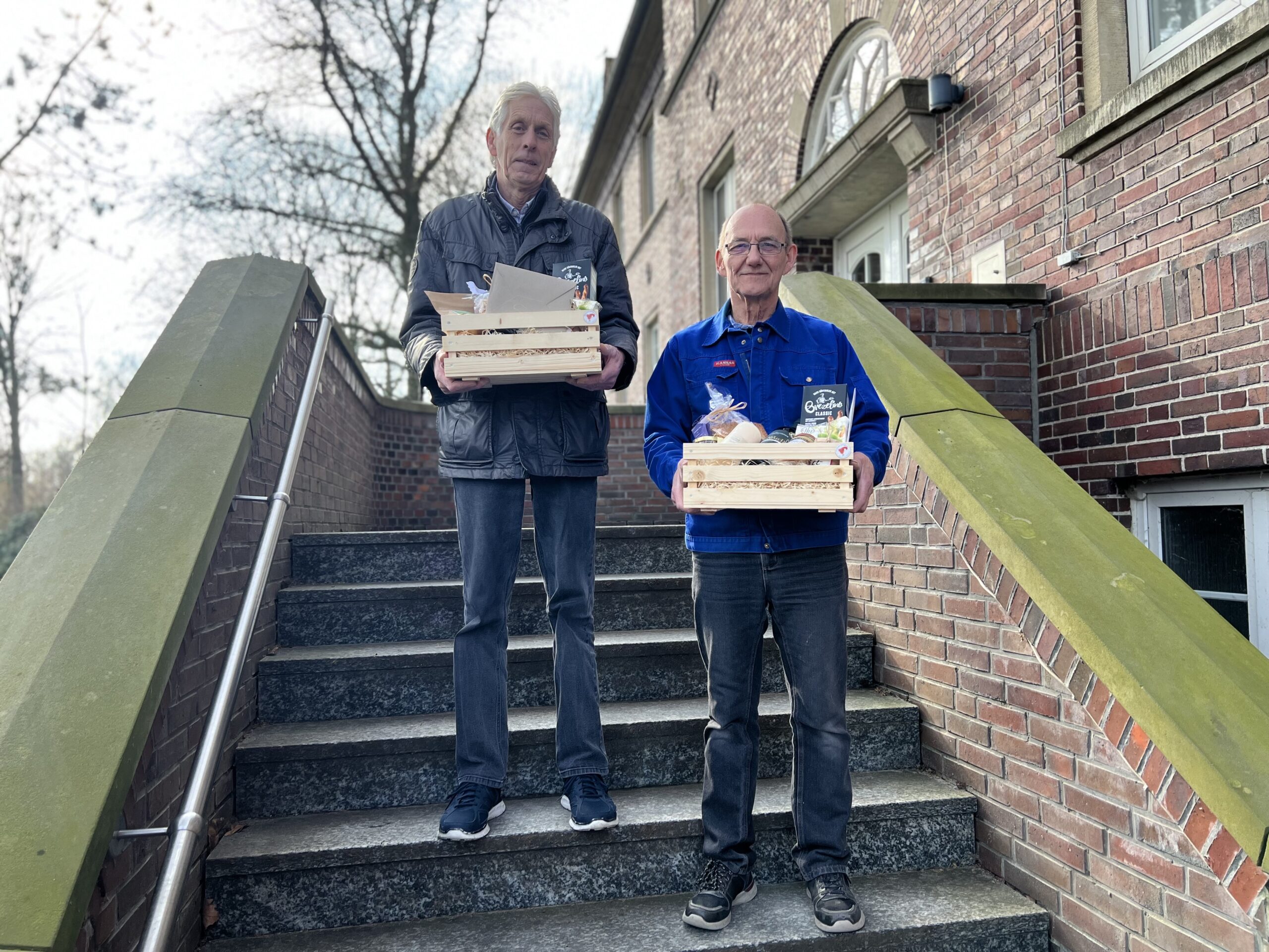 Zwei Gäste stehen draußen auf einer Treppe vor der Kamera. Sie haben jeweils ein Präsentkorb vom GPS Laden Nordseefreundlich.