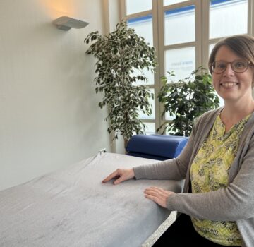 Eine junge Frau ist in einem Raum. Sie lächelt und steht neben einer Liege.