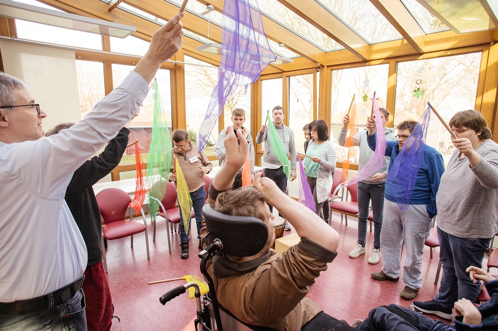 Eine Gruppe Menschen mit Behinderungen in einem Raum. Sie stehen schwingen einen Holzstock mit farbigen dünnen Tüchern hin und her.