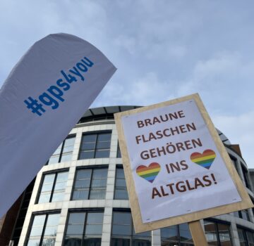 Draußen bei einer Demonstration gegen Ausgrenzung und Rechtsradikalismus vor einem Gebäude. Man sieht eine "gps4you" Flagge und ein Plakatschild mit der Aufschrift "Braune Flaschen gehören ins Altglas"