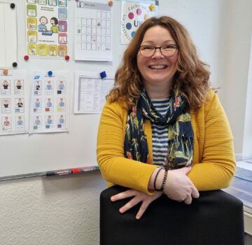 Eine GPS Mitarbeitern von der Abteilung "Unterstütze Kommunikation" in einem Raum. Sie lächelt in die Kamera. Im Hintergrund sieht man eine Flip-Chart mit vielen ängehängten Dingen.