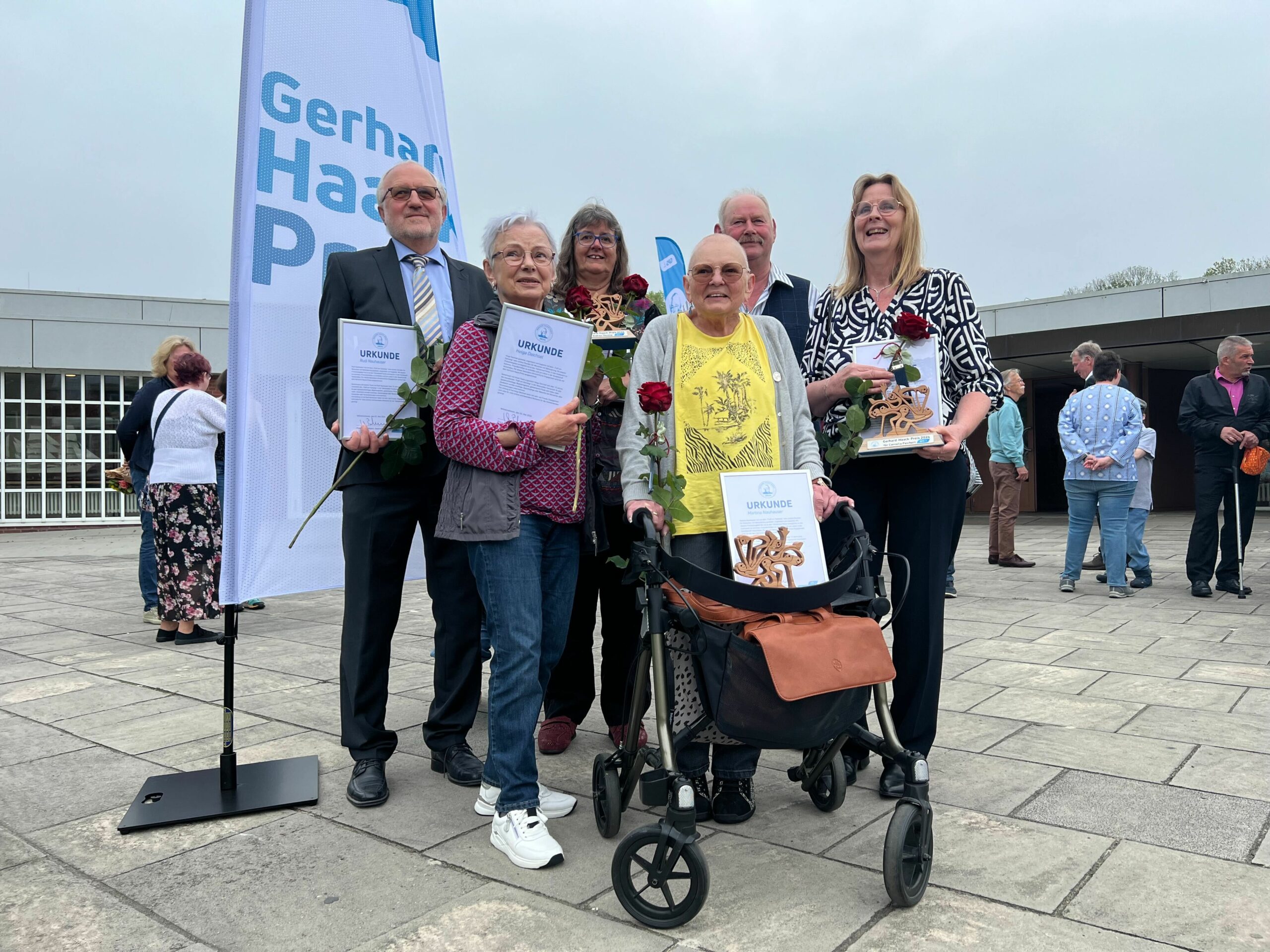 GPS Gerhard Haack Preis Nominierte sind draußen auf einem Hof und halten Ihre Urkunde in den Händen. Sie lächeln. Man sieht auch neben der Gruppe eine GPS Gerhard Haack Preis Flagge.