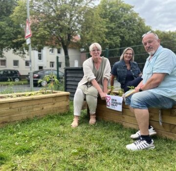 Drei GPS Mitarbeiter und Mitarbeiterinnen vom Familienzentrum Nord draußen im Garten bei sonnigen Wetter. Sie werben für eine Stiftung "Förderung Stiftung Dein Zuhause hilft.
