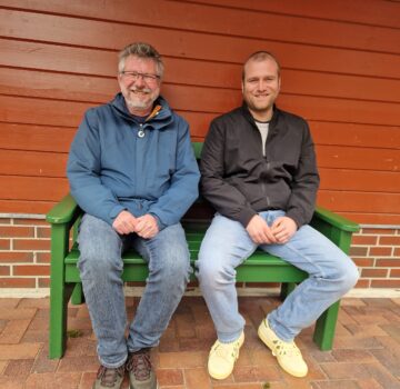 Zwei GPS Mitarbeiter sitzen auf einer Bank vor der Wand eines Hauses. Sie lächeln freundlich in die Kamera.