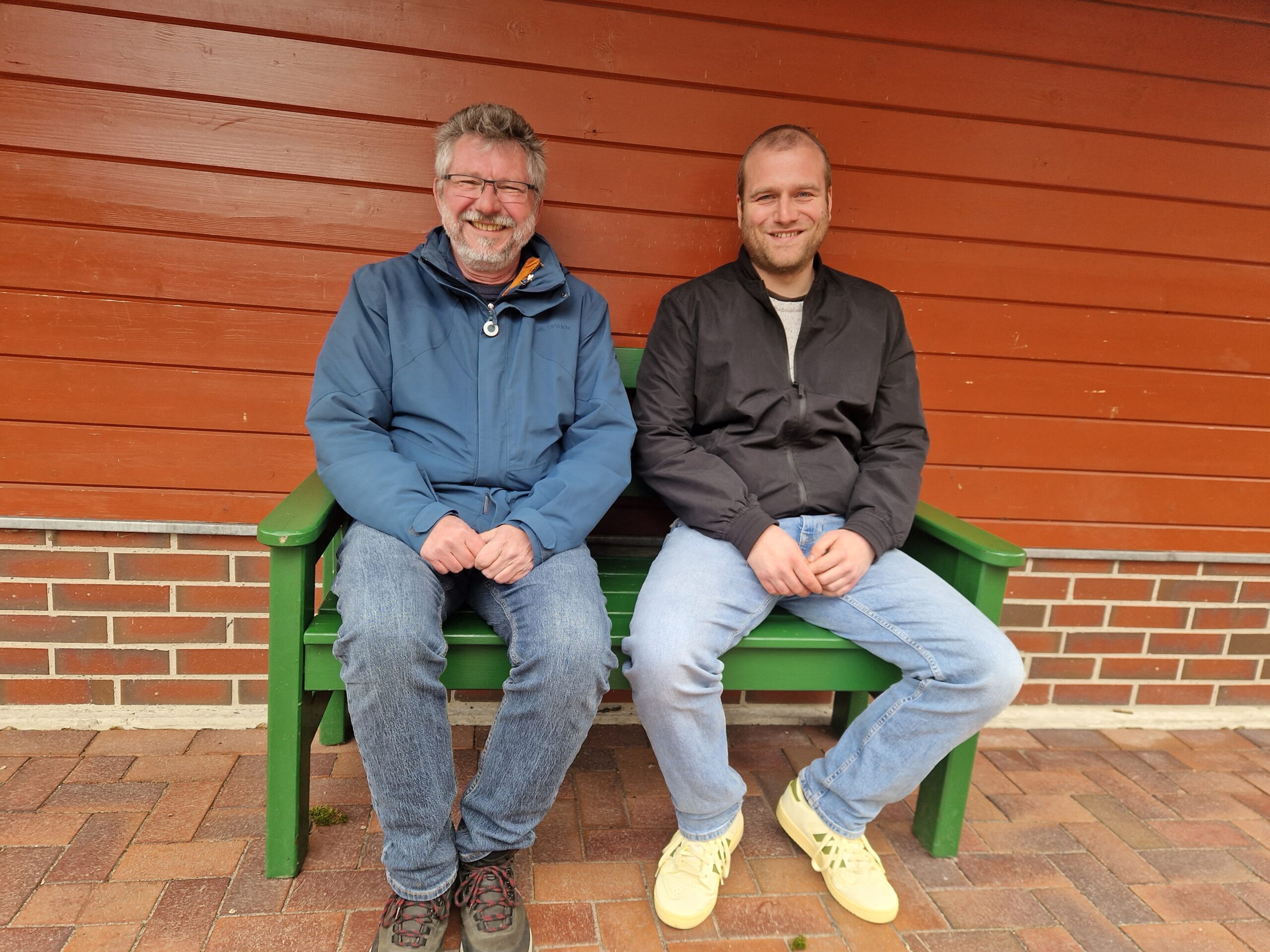 Zwei GPS Mitarbeiter sitzen auf einer Bank vor der Wand eines Hauses. Sie lächeln freundlich in die Kamera.